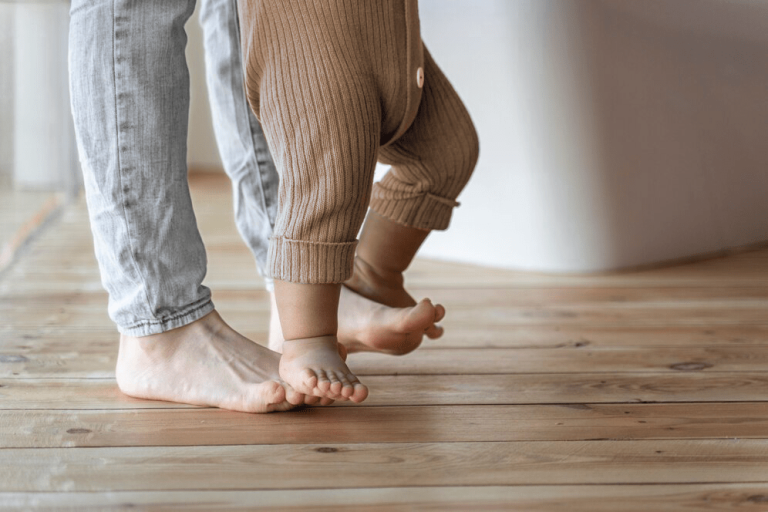 De baby staat op de voeten van vader en leert eerste stapjes zetten.