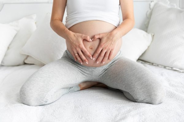 Handen op zwangere buik zittend op bed. Wanneer een buikbeeld laten maken?